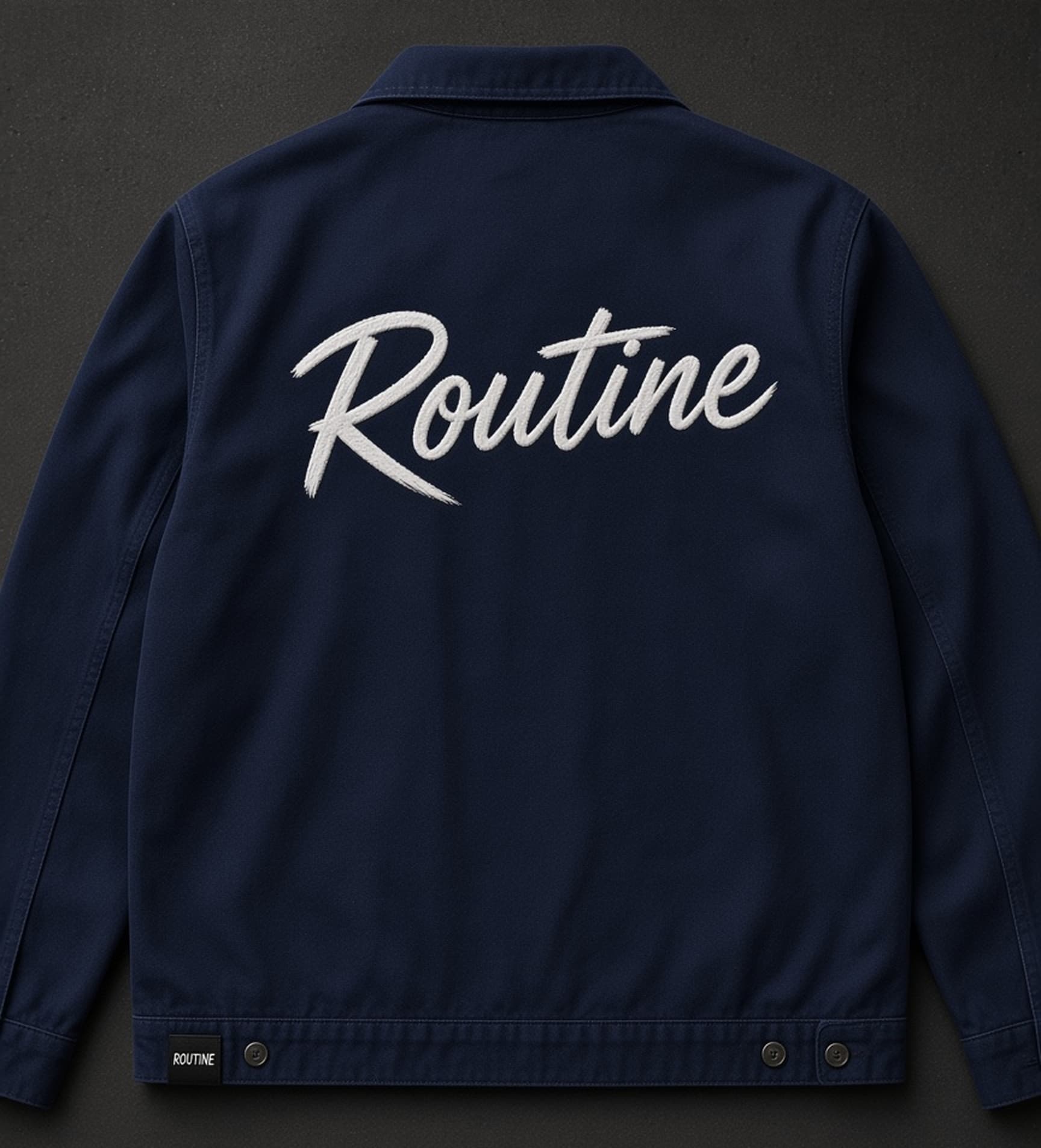 Navy work jacket laid flat, back view. Large white brush script ROUTINE across the upper back and a black ROUTINE hem tag at the left hem.